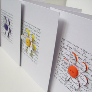 May include: Three handmade greeting cards featuring paper flowers and buttons. The cards are white with black handwritten text that reads "Happy Mother's Day".