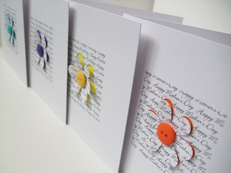 May include: Three handmade greeting cards featuring paper flowers and buttons. The cards are white with black handwritten text that reads "Happy Mother's Day".