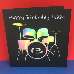 Può includere: Un biglietto nero con una batteria olografica e le parole "Happy Birthday Isaac" scritte in bianco. La batteria ha un rullante grande con il numero "13" sopra.