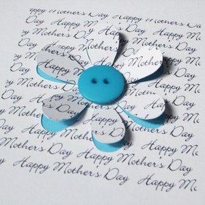 May include: A white card with the words "Happy Mother's Day" repeated many times in cursive script. A blue paper flower with a blue button in the centre is on the card.