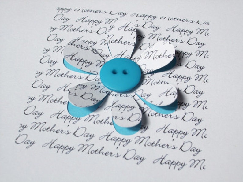 May include: A white card with the words "Happy Mother's Day" repeated many times in cursive script. A blue paper flower with a blue button in the centre is on the card.