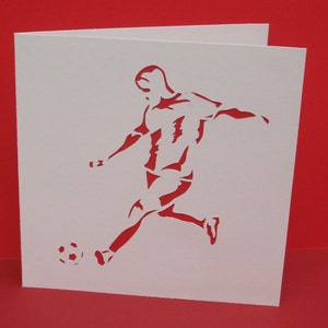May include: A white card with a silhouette of a soccer player kicking a ball. The player is in mid-air, with their leg extended and their foot about to make contact with the ball. The ball is round and has a simple design.