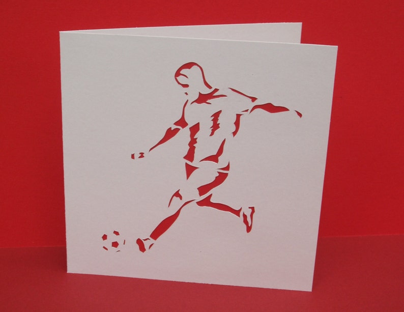 May include: A white card with a silhouette of a soccer player kicking a ball. The player is in mid-air, with their leg extended and their foot about to make contact with the ball. The ball is round and has a simple design.