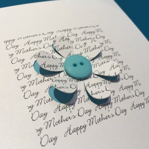 May include: A white card with a blue button in the centre of a paper flower. The flower is made of white paper with the words "Happy Mother's Day" written on each petal.