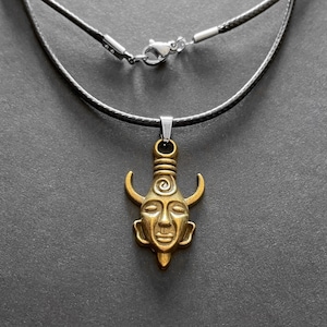 May include: A gold-toned pendant necklace with a detailed face design and horns. The necklace is on a black leather cord.