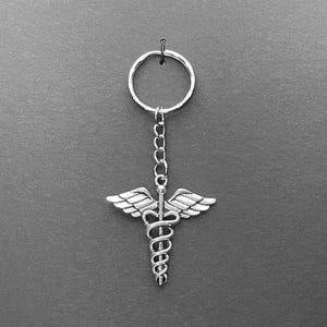 May include: Silver-colored keychain featuring the Caduceus symbol, a staff with two snakes and wings. The keychain has a ring and chain for attaching keys. The design is detailed and the metal has a shiny finish. The background is a solid gray.