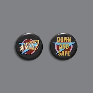 Op de afbeelding: Twee ronde zwarte knoppen met een witte ster en tekst. De eerste knop zegt "Blake's 7" in gouden letters. De tweede knop zegt "Down and Safe" in gouden letters.