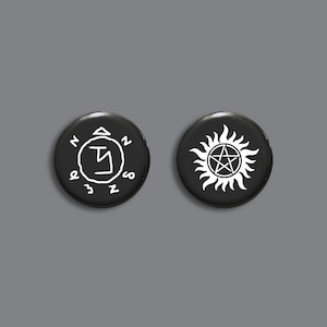 May include: Two black button badges with white designs. The first badge features a white circle with a white symbol inside. The second badge features a white pentagram with a white sunburst around it.
