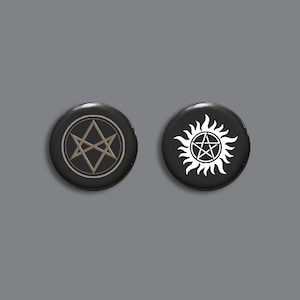 Könnte beinhalten: Zwei schwarze Knopfnadeln mit weißen und goldenen Symbolen. Die linke Nadel zeigt einen fünfzackigen Stern mit einem Dreieck im Inneren. Die rechte Nadel zeigt einen fünfzackigen Stern innerhalb eines Sonnenstrahls.