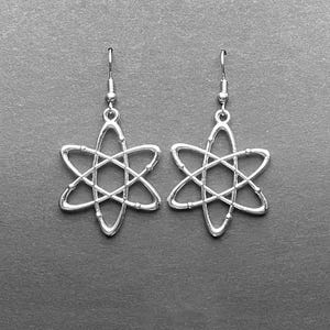 Könnte beinhalten: Ein Paar silberne Atomohrringe. Die Ohrringe sind in Form eines stilisierten Atoms mit einem zentralen Kern und umlaufenden Elektronen gestaltet.