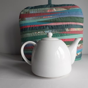 Pode incluir: Uma chaleira de cerâmica branca com tampa e alça, sentada em uma superfície branca. Atrás da chaleira, há um aquecedor de chá de tecido listrado verde, vermelho e azul.