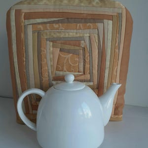 May include: A white ceramic teapot with a lid and a handle, sitting in front of a beige and brown quilted tea cozy.