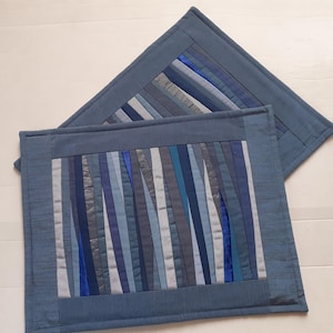 May include: Two blue quilted placemats. Each placemat has a rectangular centre with vertical strips of blue, grey, and silver fabric. The borders are a solid blue fabric. The placemats are on a white surface.