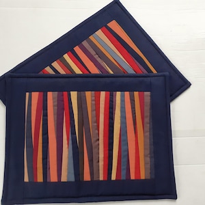 Op de afbeelding: Twee marineblauwe placemats met een kleurrijk verticaal streepontwerp. De strepen bevatten tinten rood, oranje, geel en paars. De placemats zijn rechthoekig met een vierkante bovenkant.
