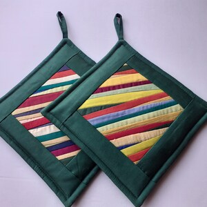 Può includere: Due presine da cucina trapuntate verdi con un inserto in tessuto a righe colorate. Le righe sono in tonalità di giallo, rosso, blu, verde e bianco.