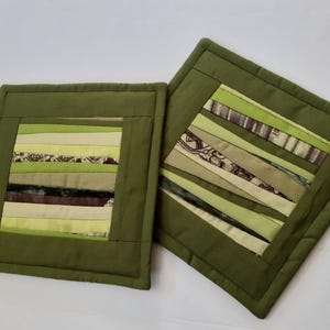 May include: Two square, quilted pot holders. The pot holders are olive green with a central patchwork design of various green and cream-coloured fabric strips. The patchwork includes patterned and solid fabrics.