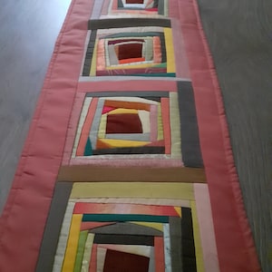 May include: A handmade table runner with a patchwork design. The runner features a series of square blocks in various colours, including pink, brown, yellow, and grey. The edges are a solid pink fabric, creating a border around the patchwork.
