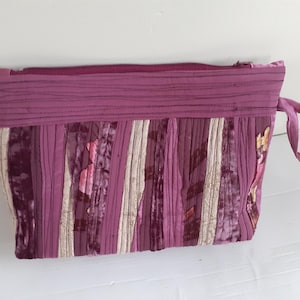 May include: A quilted, purple fabric pouch with a zipper closure and a small handle. The pouch features vertical stripes of varying shades of purple and cream-colored fabric. The top of the pouch is a solid, darker purple.