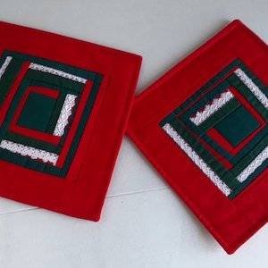 May include: Two red quilted pot holders with a green and white geometric design. The design features a square made of green fabric with white lace trim, surrounded by layers of green fabric and white lace.