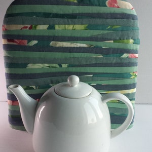 Peut inclure: Une théière en céramique blanche avec un couvercle et une poignée, posée sur un chauffe-thé en tissu rayé vert et bleu.