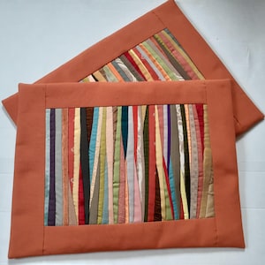 Op de afbeelding: Twee rechthoekige placemats met een verbrande oranje rand. Het midden van elke mat heeft verticale stroken in verschillende kleuren, waaronder rood, groen en goud. De placemats liggen op een wit oppervlak.