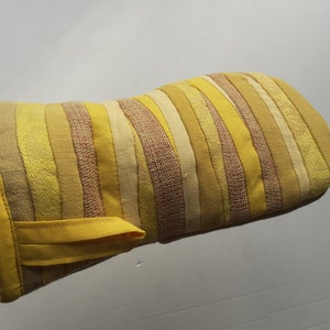 Può includere: Un guanto da forno a strisce gialle con un anello di tessuto giallo. Il guanto è realizzato con diverse tonalità di tessuto giallo cucite insieme in strisce orizzontali.