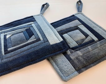 May include: Two square denim pot holders with a patchwork design. The pot holders are made from various shades of blue denim, arranged in a geometric pattern. Each pot holder has a loop for hanging.