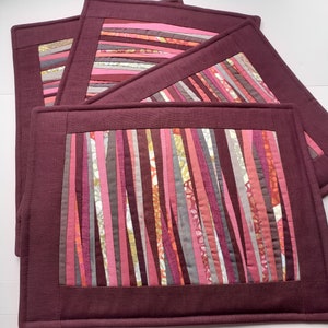 May include: Set of four quilted placemats in a burgundy color with a striped pattern of pink, orange, and gray fabric strips.
