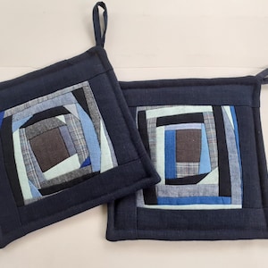 May include: Two square potholders featuring a log cabin quilt pattern in shades of navy, light blue, grey, and black. Each potholder has a solid navy blue border and a fabric loop attached to one corner for hanging, constructed from various fabric strips.