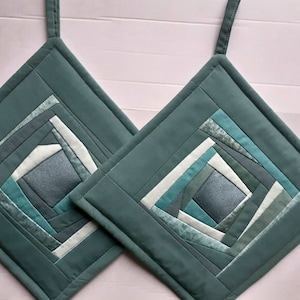 Peut inclure: Deux maniques de cuisine matelassées bleu sarcelle avec un motif géométrique de carrés sarcelle, bleus et blancs. Les maniques ont une boucle pour les suspendre.