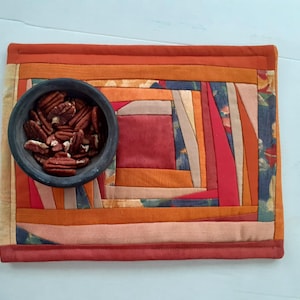 May include: A rectangular fabric placemat featuring a patchwork design of various orange, red, and brown fabric strips arranged in a log cabin pattern. A dark gray circular bowl filled with shelled pecan halves sits on the left side of the mat.