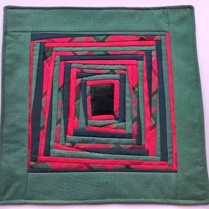 Pode incluir: Um almofada quadrado de tecido verde com um design de quadrados concêntricos em tons de rosa, verde e azul.