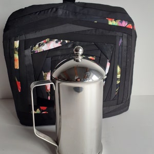 Könnte beinhalten: Eine silberne French Press Kaffeemaschine mit einer schwarzen Kaffeekanne aus Stoff mit floralem Muster. Die Kanne hat einen Griff und ist gesteppt.