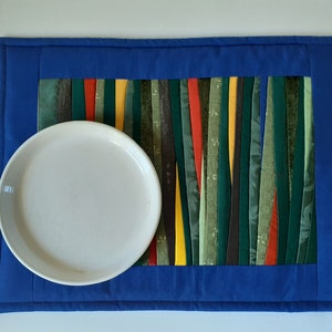 May include: A blue quilted placemat with a colorful striped design in the center. A white plate is placed on top of the placemat.