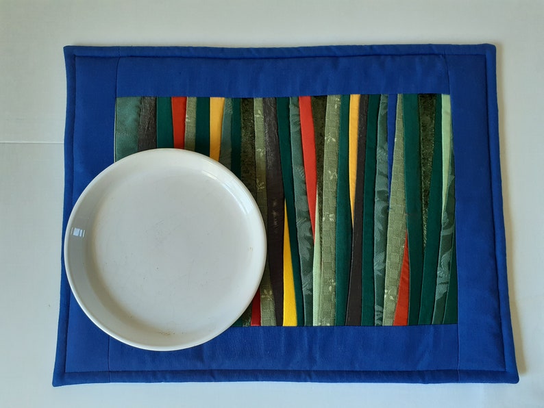 May include: A blue quilted placemat with a colorful striped design in the center. A white plate is placed on top of the placemat.