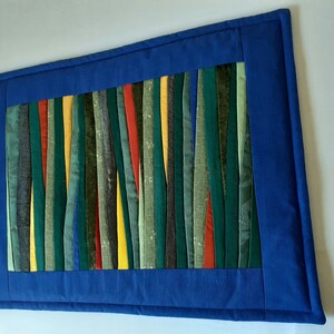 May include: A blue fabric wall hanging with a colorful striped design. The stripes are made of different fabrics and colors, including green, yellow, red, and brown.