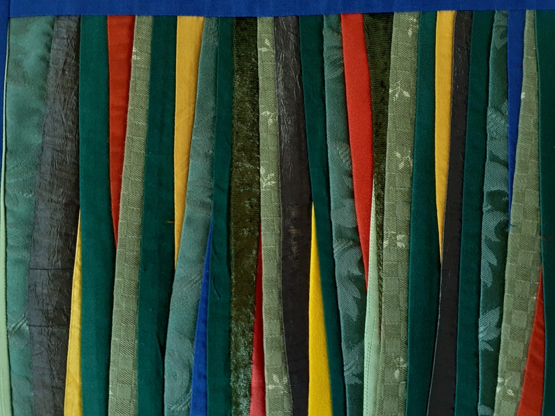 May include: A close-up of a fabric wall hanging made of strips of fabric in various shades of green, blue, yellow, and red. The fabric strips are sewn together to create a vertical pattern.