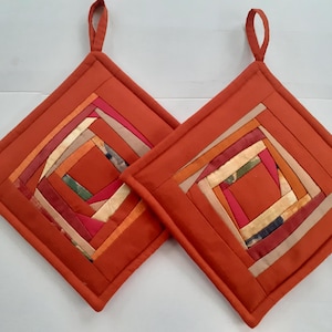 Op de afbeelding: Twee vierkante pannenlappen met een log cabin quiltpatroon in tinten oranje, roze en beige. Elke pannenlap heeft een effen oranje rand en een stoffen lus aan de bovenhoek om op te hangen. Het ontwerp bestaat uit verschillende stroken stof.