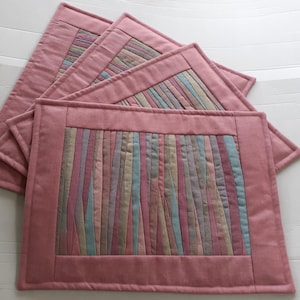 Peut inclure: Un ensemble de quatre sets de table rectangulaires en tissu avec une bordure rose poudré unie entourant un panneau central composé de bandes verticales multicolores dans des tons de rose, bleu, beige et gris, avec des coutures matelassées.