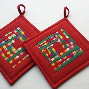 May include: Two red quilted pot holders with a colorful patchwork design in the center. The pot holders have a loop for hanging.