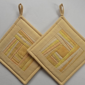 May include: Two square, quilted pot holders in shades of beige and yellow. Each has a geometric design with a loop for hanging. The design features a patchwork of various yellow and beige fabrics. These kitchen accessories are ideal for handling hot cookware.