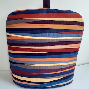 Puede incluir: Un cubreteiera acolchado con asa, con bandas horizontales de varios colores, incluyendo azul marino, rojo, naranja y dorado. El cubreteiera tiene una parte superior redondeada y un asa rectangular de tela morada oscura.