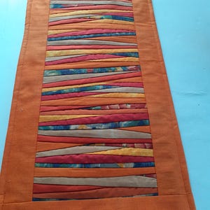 Könnte beinhalten: Ein rechteckiger, orangefarbener Tischläufer mit einem zentralen Feld aus farbenfrohen, horizontalen Stoffstreifen. Die Streifen sind in Orange-, Rot-, Blau- und Beigetönen gehalten. Der Läufer hat einen durchgehenden orangefarbenen Rand.