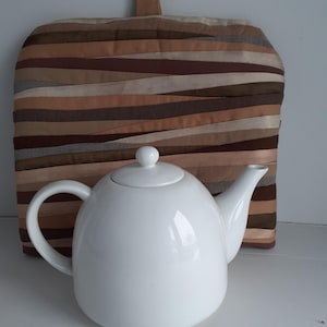 May include: A white ceramic teapot with a curved handle and spout, set in front of a tea cozy. The tea cozy is made of horizontal strips of fabric in shades of brown, beige, and tan. The teapot has a rounded shape and a small lid.