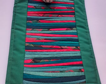 May include: A green quilted table runner with a colorful striped pattern. A white ceramic cup with blue and white designs sits on top of the runner.