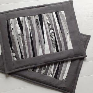 May include: Two rectangular fabric placemats featuring a central panel of vertical strips in shades of black, white, and gray. The center is framed by a solid dark gray border with visible stitching. The fabric has a textured, quilted appearance.