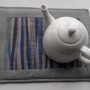 May include: A white teapot sits on a grey and blue striped fabric coaster. The coaster has a grey border.