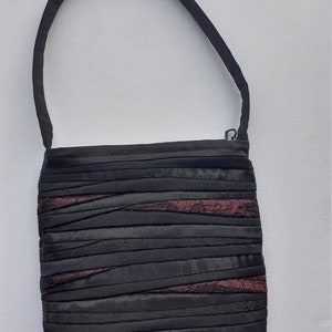Peut inclure: Sac à bandoulière noir avec une longue sangle. Le corps du sac est composé de bandes horizontales de tissu noir, avec des sections de tissu bordeaux tissées. Le sac a une fermeture éclair.