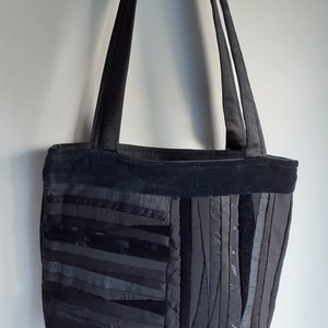 Może przedstawiać: Czarna torba typu tote wykonana z pasków materiału. Torba ma szeroką, płaską podstawę i dwa długie paski.