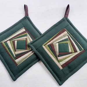 Op de afbeelding: Twee groene, gestikte ovenwanten met een geometrisch patroon van vierkanten in tinten groen, bruin en crème. De ovenwanten hebben een lusvormige handgreep.